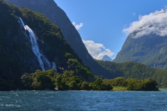 Majestic waterfall flows from forested mountain into the water under a clear blue sky, summer,