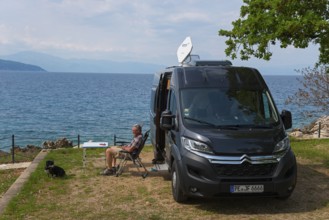 A campsite by the sea, a man sits next to a motorhome and enjoys the view with a dog, Glavotok, Krk