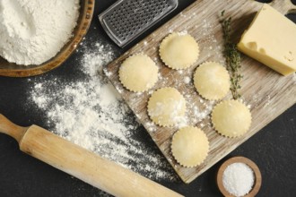 Fresh pasta dough is rolled out on a wooden board surrounded by flour, cheese, and herbs. Round