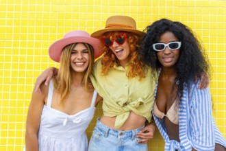 Three cheerful diverse women with colorful hats and sunglasses are posing together against a bright