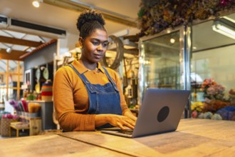 Young florist managing online orders using laptop in her flower shop, surrounded by colorful
