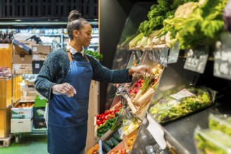 Grocery store employee wearing gloves arranging organic vegetables on display in supermarket