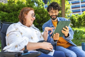 Man and a woman are using a digital tablet in a park, she is sitting in a wheelchair and they are