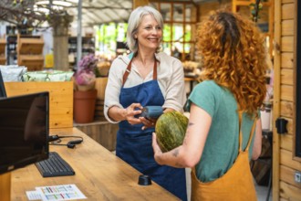 Smiling greengrocer holding pos terminal while customer paying with contactless payment for