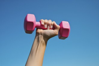 Woman's hand holding pink dumbbell free weight in front of blue sky. Generative Ai, AI generated