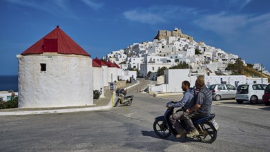 Two people on motorbike in front of white village and windmills on hill, Querini Castle, Chora,