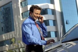 Young Indian Executive with tie working on laptop and cell phone on car bonnet in front of modern
