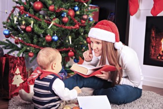 Toddler boy and his mother reading a book in front of christmas tree