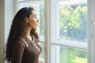 A young woman with long, curly hair is looking out a large window. Bright sunlight fills the room,