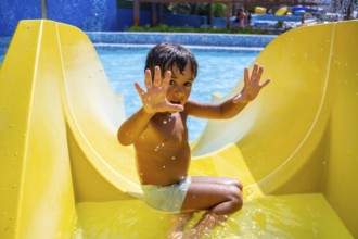 Happy child sliding down yellow water slide in amusement park, having fun during summer vacation