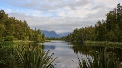 A tranquil lake surrounded by lush vegetation and mountains, with a slightly cloudy sky, summer,