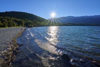 A tranquil lake with sunbeams hitting the water and shore, surrounded by forested mountains under