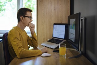 A middle-aged man sits at a wooden desk in a home office, focused on a computer screen displaying