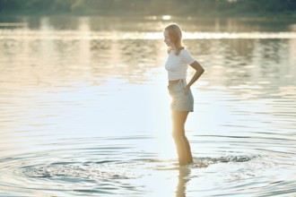 A young woman in a white top and denim shorts stands in shallow water during sunset. The golden