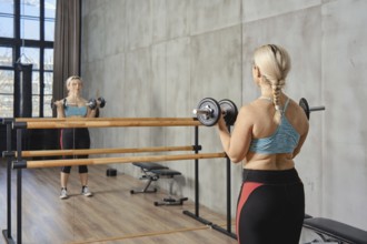 A woman with a braid exercises with dumbbells in a contemporary gym. She is focused, using a mirror