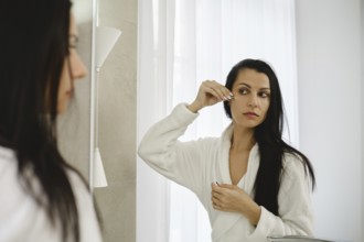 A woman in a white robe looks into a mirror while applying a beauty treatment to her under eye skin