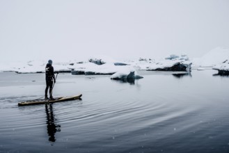 Man stand up paddling, snowfall, glacial lake Jökulsarlon at Vatnajökull, Iceland* Man stand up