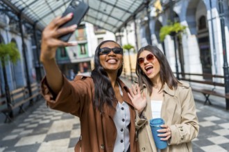 Two cheerful businesswomen taking a selfie with a smartphone and waving at the camera while walking