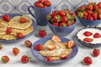 Pancakes sprinkled with icing sugar and fresh strawberries on plates next to yoghurt, blue crockery