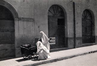 Two Arab women with prams, 1910s, A veiled person sits next to a pram in front of an old building