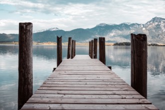 A wooden footbridge leads into a tranquil lake, surrounded by mountains under an open sky