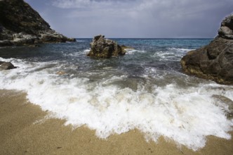 Energetic waves meet rocks on the sandy beach under a slightly cloudy sky, Ricadi, Capo Vaticano
