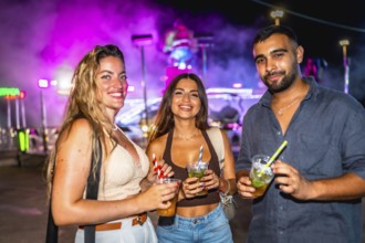 Three friends are holding cocktails while enjoying the vibrant nightlife at an amusement park,