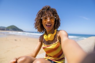 Happy young woman with headphones and sunglasses taking a selfie on a sunny beach, enjoying her
