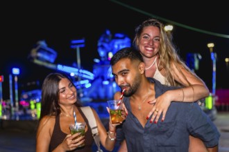Three cheerful friends enjoying refreshing cocktails at an amusement park, illuminated by vibrant