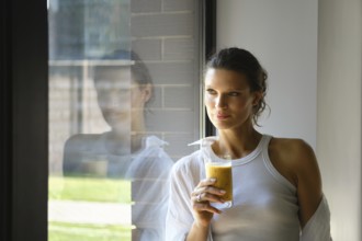 In a stylish, well-lit room, a woman holds a glass of fresh juice. She gazes thoughtfully out the