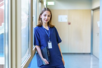 Confident young doctor wearing blue scrubs holding a clipboard while standing in a modern hospital