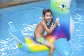 Smiling boy riding a colorful inflatable sea lion toy while splashing in the swimming pool during a