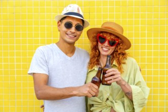 Happy couple toasting with beer bottles while enjoying a summer day against a vibrant yellow tiled