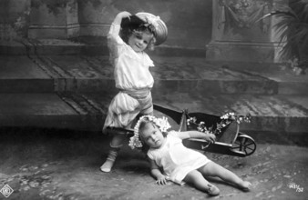 Child falls out of wheelbarrow, 1910, Two children playing in the garden, one with a wreath of