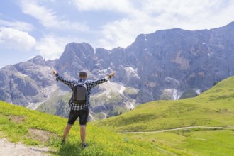 A hiker with raised arms looks at an impressive mountain scenery in a meadow, Alpe di Siusi,