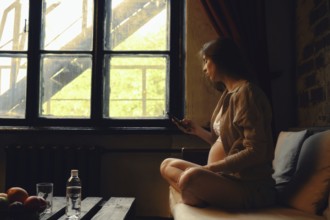 A pregnant woman sits cross-legged on a couch in a cozy room, looking out of the window. She is