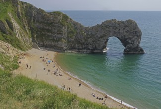 People relaxing on the beach in front of a striking arched cliff and blue sea, Durdle Door,