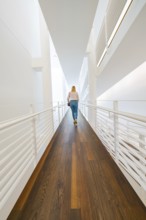 Woman walking along a modern corridor characterised by light-coloured walls and wooden flooring,