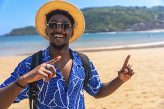 Happy black tourist smiling widely while pointing both index fingers up at a beautiful beach,