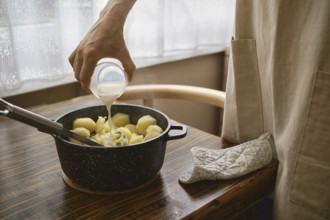 A hand pours warm milk onto a pot filled with boiled potatoes on a wooden table. The process of
