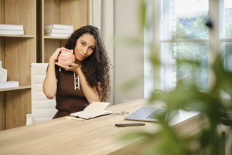In a contemporary office space, a woman with curly hair sits at a wooden desk, smiling as she holds