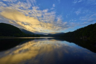 The dramatic dawn is reflected on the calm lake, surrounded by rolling hills and mountains, summer,