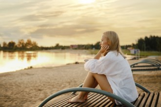A woman sits comfortably on a wooden bench beside a serene lake, gazing at the vibrant sunset. The