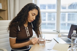 A woman with curly hair sits at a desk in a contemporary office, holding a pill and a glass of