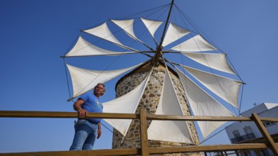 A man leaning against a wooden fence in front of a windmill with white sails and a building in the
