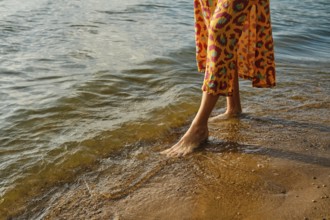 Unrecognizable woman walks barefoot along a sandy beach as waves gently lap at her feet. She wears