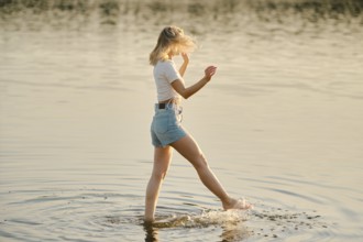 A woman walks through shallow water, splashing lightly as golden sunlight reflects off the surface.