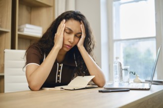 Tired young woman sits at her desk, visibly stressed and holding her head in her hands. The