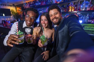 Three friends smiling and capturing a selfie while enjoying colorful cocktails at a lively bar,