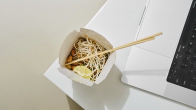 A cardboard bowl of noodles with vegetables and lime sits next to a laptop on a white table. The